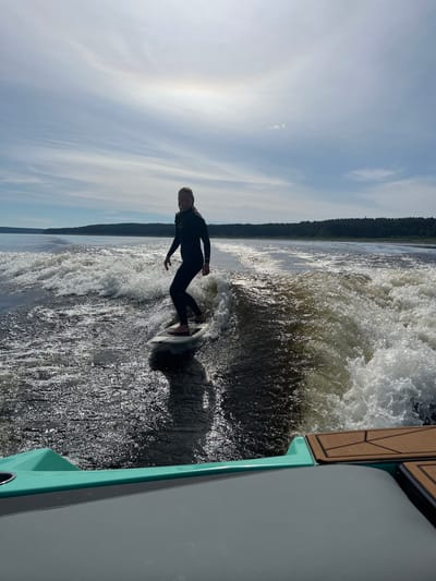 A person wearing a black wetsuit surfs on a wakeboard behind a boat. The person is positioned on the right side of the wakeboard, with their left foot forward. The wakeboarder is in mid-action, riding the crest of a wave. The water is dark and choppy, with a significant amount of white foam from the boat's wake. The background shows a dense, dark green forest line on a distant shore. Above the treeline, a pale blue sky with scattered white clouds is visible. A sun halo is present in the upper portion of the sky, indicating bright sunlight. The foreground shows part of the boat's interior, with a teal-colored edge and grey seating. The image appears to be taken from the perspective of someone on the boat, looking back at the wakeboarder. The location is the city of Gromovo, Russia.