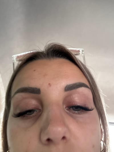 The image is a close-up, low-angle, selfie-style shot of a young woman's face, primarily focusing on her eyes and forehead. The lighting appears to be natural, possibly from a window or overhead source, casting soft shadows. She has dark, defined eyebrows, and her eyelids are reddish, suggesting possible irritation or a cosmetic procedure like eyelash extensions, which are visible on her lashes. There is a small mole on her forehead. She is wearing hoop earrings. The background is out of focus, but hints at an indoor environment with what might be a car ceiling or a similar textured surface above her. The woman is looking directly at the camera. The image does not contain any information about the city of Ust'-Isha, Russia.