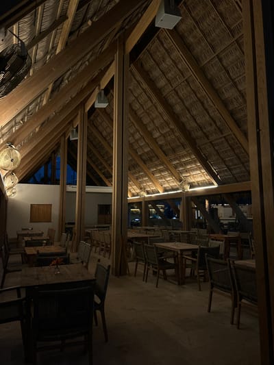 This image depicts the interior of a spacious, open-air or semi-open-air restaurant or bar, likely a palapa structure, at night. The dominant architectural features are a high, pitched thatched roof made of natural fibers and a sturdy framework of large wooden beams. These beams run both diagonally and horizontally, creating a rustic and tropical aesthetic.
Artificial lighting plays a key role in illuminating the space. A strip of warm light runs along one of the prominent wooden beams. Several smaller, round fans with integrated lights are mounted to the ceiling structure, while a larger, caged industrial-style fan is visible in the foreground, also attached to the wooden beams. Rectangular grey speakers, one bearing the "JBL" logo, are mounted on other beams.
The dining area below consists of numerous wooden tables and chairs. The chairs feature woven seats and backs, complementing the natural materials of the roof. The tables are mostly rectangular. The floor appears to be a light-colored, possibly concrete or tiled, surface. No people are present, giving the scene a quiet, serene atmosphere, perhaps indicating after-hours or a lull in activity.
In the background, through openings in the structure, parts of an adjacent building or complex are visible, including white walls and windows with closed wooden shutters. A dark night sky and distant blue lights suggest other outdoor areas or pools, reinforcing the impression of a resort or coastal establishment. The overall ambiance is characteristic of a tropical dining venue in Mazatlán, Mexico, during the evening.