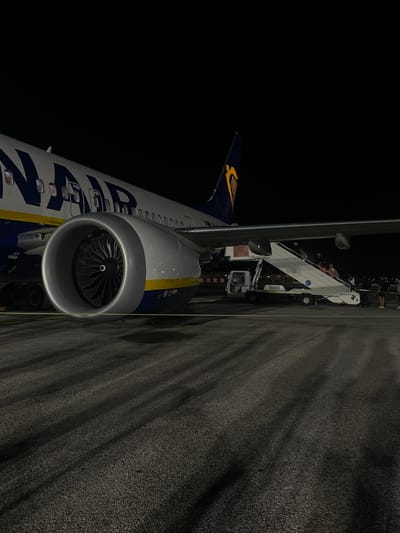 The image captures a Ryanair aircraft on an airport tarmac at night, with passengers either boarding or disembarking via mobile stairs. The scene is illuminated by artificial lighting against a dark sky, suggesting it was captured at an airport in Pisa, Italy.

In the foreground and mid-ground, the large turbofan engine of the aircraft is prominently featured, revealing its intake fan blades. The left side of the frame shows the aircraft's fuselage, painted in Ryanair's distinctive blue and white livery, with parts of the "RYANAIR" text visible. Further back, the blue tail fin with the airline's harp logo can be seen against the dark expanse of the night sky.

A mobile boarding stair is positioned next to the aircraft's door, and several people, appearing to be passengers, are visible on the stairs, engaged in the boarding or disembarking process. The tarmac on the right side of the image is dark and textured, showing patterns of light and shadow cast by the artificial illumination. The overall impression is that of an active airport environment during nighttime operations.