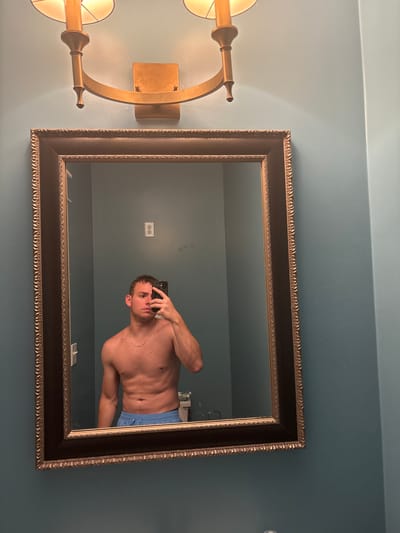 The image shows a person taking a mirror selfie in a bathroom. The person is a shirtless male with muscular build, wearing blue boxer briefs. He is holding a smartphone in his right hand, angled to capture his reflection. The bathroom has teal-colored walls and a large, ornate mirror with a dark frame. To the left of the mirror, there is a golden wall sconce with two lights. The lighting appears to be artificial, suggesting it could be nighttime or an indoor setting with the lights on. The overall mood is casual and self-observational. No visible text is present in the image. The location context of Panama City Beach, USA, is provided, which could imply the setting is a residential bathroom or hotel.