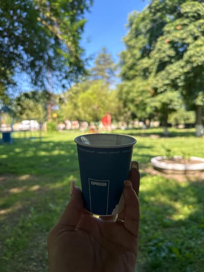 The image shows a close-up of a hand holding a blue paper cup of espresso in an outdoor park setting. The cup has the word "ESPRESSO" printed vertically on it in white text, along with some smaller text that reads "express yourself". The hand has neatly manicured nails with light pink polish and is also wearing a gold ring on one finger.
The background is a blur of green grass, trees, and a bright blue sky, suggesting a sunny day. In the mid-ground, there are hints of playground equipment and possibly some vehicles, suggesting this is a public park within a town or city. The overall impression is one of a relaxed moment enjoying a coffee outdoors. The location is identified as Salmanovo, Bulgaria.
