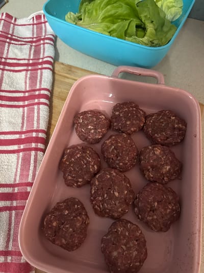 The image captures a top-down view of food preparation taking place on a kitchen counter. In the center, a light pink, rectangular ceramic baking dish with handles contains eleven irregularly shaped, raw meat patties or meatballs. These appear to be made from ground meat, mixed with lighter-colored ingredients, possibly breadcrumbs or oats.
To the left, a light blue plastic container holds fresh, vibrant green lettuce leaves, some with visible water droplets, suggesting they have just been washed. The containers rest on a light-colored countertop, with a wooden cutting board partially visible beneath the pink dish. In the bottom foreground, a red and white checkered kitchen towel or cloth is spread out.
The scene depicts ingredients ready for a meal, likely hamburgers or a similar dish involving ground meat and fresh greens. No people are visible in the image, nor is any text. The lighting suggests an indoor setting, likely during the daytime. The provided context indicates the location is Subotica, Serbia, though no specific visual cues in the image confirm this.