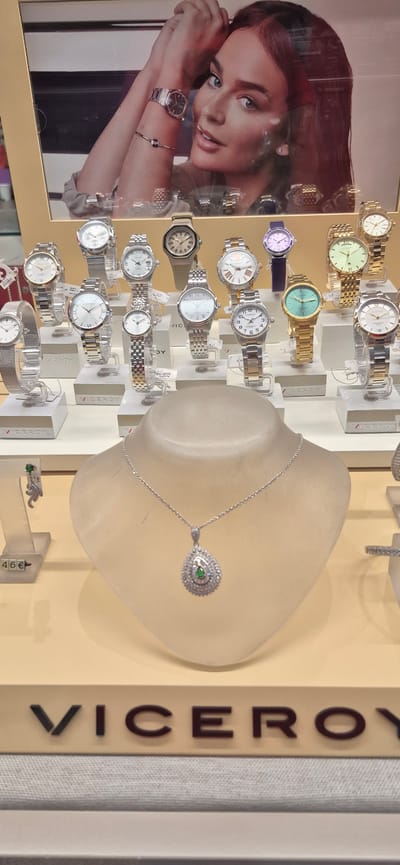 The image shows a display of Viceroy brand watches and jewelry in a store located at Rua da Eça de Queirós, 23, 4500-308 Póvoa de Varzim, Portugal. The display features multiple watches of various styles and colors, arranged on white stands. In the foreground, a mannequin bust showcases a silver necklace with a teardrop-shaped pendant containing a green stone. To the left, a price tag displays "46€" next to a pair of dangling earrings with a green stone. A large advertisement poster of a woman wearing a watch is visible at the top of the display. The overall lighting is bright, highlighting the merchandise.