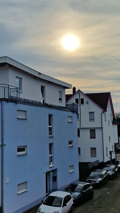 The image depicts a street scene in Villingen-Schwenningen, Germany, characterized by residential buildings and parked cars. The sun is setting, casting a warm glow across the sky and illuminating the cloudy atmosphere. The buildings, varying in color from white to light blue, have multiple windows and balconies. A metallic fire escape is visible on one of the buildings. Several cars are parked in a row along the street, including a white car with black accents prominently featured in the foreground. The overall mood is calm and serene, typical of an evening in a suburban or urban residential area. No people are visible in the image. There is no visible text.