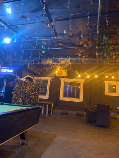 This image depicts an outdoor or semi-outdoor covered area within a commercial establishment in Igbessa, Nigeria, illuminated at night. The scene is characterized by two distinct lighting zones.
In the foreground left, a dark-framed billiard table with a green felt surface is visible. The lower legs and feet of an individual, wearing light-colored sandals, are positioned underneath the table, suggesting a person is standing nearby. To the left of the billiard table, a densely covered artificial foliage screen stands next to a structure where a partially visible "RECEPTION" sign, illuminated in blue, is present. A bright blue light fixture hangs from the ceiling in this area.
The overhead structure consists of a dark metal framework from which various decorative elements are suspended. These include white string lights, artificial green and autumn-colored leaves/vines, and additional string lights with warm, yellow-glowing bulbs.
The background features a dark-colored building wall with several white-framed windows of varying sizes. Above the central windows, an outdoor air conditioning unit is mounted on the wall. Below these windows, two dark wicker-style armchairs are arranged around a small, dark table. Another small, light-colored table with a pale wooden top and legs is situated between the billiard table and the building wall. The floor appears to be paved with dark tiles or concrete.