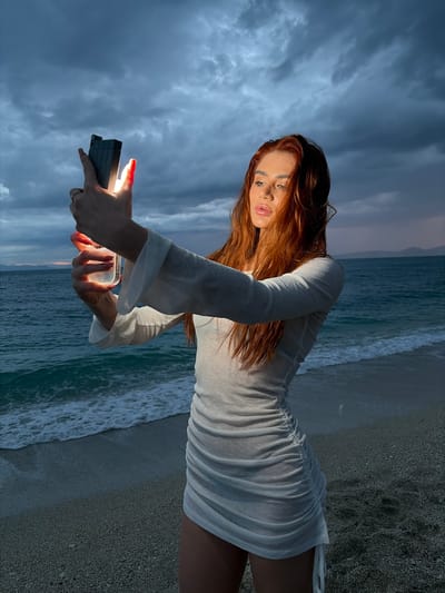 A woman with red hair and fair skin, wearing a short white dress with long sleeves, is standing on a beach. The beach is near the city of Alimos, Greece, as the location data states. She is holding a black mobile phone in her hands, and light is emanating from the screen. The background includes a cloudy sky and the sea. The photograph appears to be taken at dusk or dawn, given the lighting. Waves are visible along the shoreline.