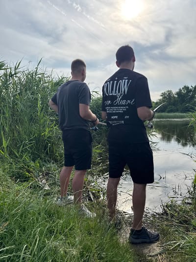 The image depicts two adult males fishing from the grassy bank of a body of water in the city of Zamfir, Bulgaria. The sky above is partly cloudy with a bright sun visible in the upper right quadrant, casting reflections on the water's surface. The immediate foreground and banks are covered with dense, tall green reeds and grass, with a line of trees visible in the distance across the water.

The man on the left, seen from behind, wears a dark gray short-sleeved t-shirt, dark shorts, and white sneakers. He holds a fishing rod with a reel in his hands, positioned to his right side. The man on the right, also viewed from behind, wears a black short-sleeved t-shirt that displays white text and graphics, including "ESTD 1987," "FOLLOW YOUR HEART," "HOPE," and "JOY." He is dressed in black shorts and dark athletic shoes with blue accents. He holds a fishing rod extending over the water, with the fishing line visible entering the water. Both individuals are standing near the water's edge, facing away from the viewer and toward the body of water. Scattered light-colored debris is visible among the tall grass near the feet of the man on the left.