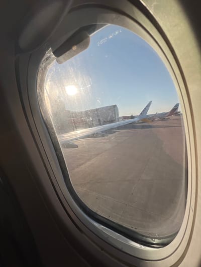 The image captures an external view from a commercial aircraft passenger window, specifically located at Palma de Mallorca Airport (PMI) in Son Santjoan, Spain. The oval window frame and a portion of the viewing aircraft's wing are prominent in the foreground. The wing extends from the lower left towards the mid-right, reflecting bright sunlight. Beyond the wing, a paved airport tarmac is visible. In the mid-ground, the tail section of a white aircraft with "RYANAIR" branding in blue and yellow is parked on the tarmac. To its right, the vertical stabilizer of another aircraft, displaying red and white livery consistent with Eurowings, is partially visible. In the background, a large, grey airport building or jet bridge is positioned to the left. The sky is clear and blue, with the sun producing a strong glare and lens flare in the upper left quadrant of the window, indicating either morning or late afternoon. No individuals are visible in the scene.