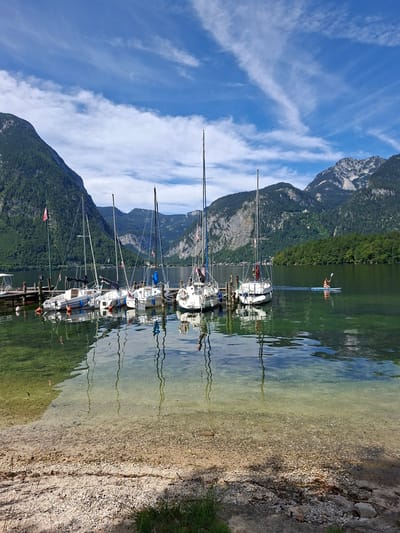 A wide shot of a lake with sailboats docked at a pier on the left and a person paddleboarding on the right. The lake is clear and the bottom is visible in the foreground. Mountains covered in trees rise up from the far side of the lake and in the background. The sky is blue with white clouds. The location is the Traunsee lake in Obertraun, Austria.