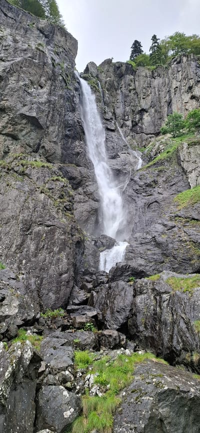 This is a full-shot, eye-level, vertical image of a tall, rocky waterfall. The waterfall is situated within a rocky canyon with lush green grass growing in patches on the rocks. Trees are visible at the top of the canyon, and the sky is overcast. The water cascades down a sheer rock face, creating a powerful and dynamic effect. The rocks are dark gray and appear wet, suggesting recent rain or mist from the waterfall. The overall impression is one of natural beauty and ruggedness. There are no people or animals visible in the image.