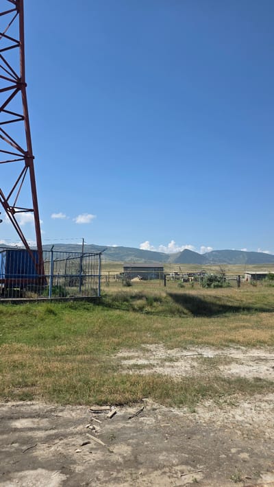 The image shows a rural landscape under a clear blue sky with scattered white clouds. In the foreground on the left, a portion of a large red metal structure, possibly a tower or support, is visible. Adjacent to this structure is a fenced enclosure with a blue tarp-covered object inside. The fence is made of metal bars and posts. Beyond the fence, there is a field of dry and green grass. In the middle ground, several low-rise buildings, appearing to be simple structures or sheds, are scattered across the landscape. A yellow pipe runs horizontally across the scene in front of these buildings. In the background, rolling hills and mountains form a natural horizon. The ground in the immediate foreground is dirt and appears somewhat disturbed, with scattered pieces of wood and debris. The overall scene suggests a remote, possibly agricultural or industrial area outside of Novyy Chirkey, Russia. There are no people visible in the image.