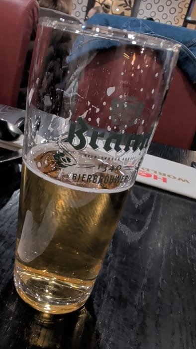 The image depicts a clear, tall glass, approximately three-quarters full of a light amber liquid consistent with beer, positioned centrally on a dark, grain-patterned wooden table. The glass bears a green and white logo partially reading "Brand 1340 BIERBROUWERIJ". Small bubbles adhere to the interior surface of the glass, and a minimal layer of foam rests on the liquid. To the right of the glass, a white rectangular object with inverted red text, displaying "WORLD" and partial letters "S" and "H", is visible on the table. A metallic utensil is partially visible to the left of the glass. The background features red upholstered seating, with a person's leg clad in blue denim jeans discernible behind the glass. Further back, patterned wall decor with light circular elements on a darker background is present. The scene is an indoor establishment, likely a bar or restaurant, located in the city of Horst, Netherlands.