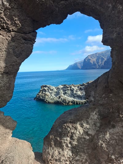 This is a photograph taken through a natural rock archway at Acantilados de Los Gigantes in Santiago del Teide, Spain. The view looks out towards the Atlantic Ocean, which is a vibrant turquoise blue. In the middle ground, a rocky outcrop or small island rises from the sea. In the background, the dramatic cliffs of Los Gigantes dominate the horizon, their rugged slopes meeting the deep blue sea under a bright, partly cloudy sky. The foreground is framed by the textured, dark brown rock of the archway, casting shadows on its rough surface. There are no people or animals visible in the image.
