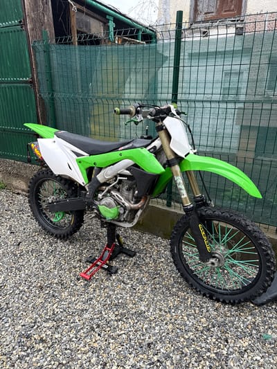 The image shows a green and white dirt bike parked on a gravel surface. The bike is oriented diagonally in the frame. A green wire fence is visible in the background behind the bike, with a building visible behind the fence. The gravel surface surrounds the bike, with a red stand supporting the bike. The location of the scene is Mikre, Bulgaria.