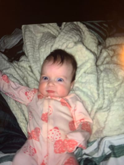 The image displays an infant lying supine on a textured, light-colored blanket. The infant has fair skin, dark brown hair, and wide-set blue eyes, with a facial expression characterized by puckered lips and a slight grimace. The infant is wearing a pink, long-sleeved, footed sleeper with a front zipper closure, patterned with light pink and darker pink hearts, some featuring white polka dots. The infant's left arm is slightly extended to the side. The blanket beneath the infant has a visible braided or ribbed texture. To the left and lower edge of the frame, dark blue or green fabric is visible, along with a faintly striped fabric section on the right. The background above the infant is dark. A subtle orange-yellow lens flare is present on the right side of the image. The precise location within the city of Tulsa, United States of America, cannot be determined from the visual information presented.