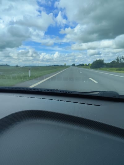 The image is a point-of-view shot from inside a vehicle, looking through the windshield at a highway. The road appears to be a multi-lane highway with white lane markings and a grassy median. The sky is partly cloudy with white and grey clouds. The foreground shows the dashboard of the car and the base of the windshield wipers. The environment suggests a rural or suburban setting with fields and trees visible in the distance. There are no people or animals visible in the image. The time of day appears to be daytime, given the lighting. The location is identified as Sokol'niki, Russia.