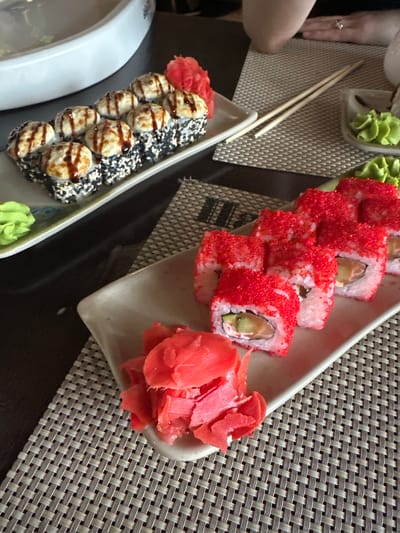 A high-angle shot shows two plates of sushi on a dark brown table. The plates are off-white and rectangular with rounded corners. One plate contains sushi with red tobiko (flying fish roe) and pickled ginger on the right. The other plate contains sushi topped with creamy sauce and black sesame seeds, accompanied by pickled ginger and wasabi. Chopsticks rest between the plates. A hand wearing a ring is visible on the left side of the frame. The table is covered with a woven placemat. This image was taken in Pokrov, Russia.