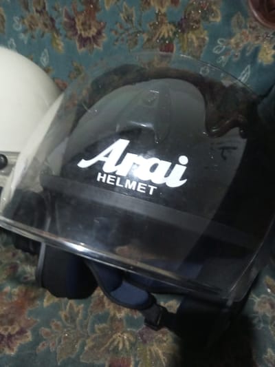 The image depicts two motorcycle helmets resting on a floral-patterned textile surface. A black Arai full-face motorcycle helmet, bearing the white "Arai HELMET" logo on its front, occupies the majority of the frame, with its clear visor lowered. To the left, a portion of a white motorcycle helmet is partially visible. The textile surface features a teal/green background adorned with interwoven floral motifs in brown, yellow, and reddish hues. The scene is observed in B. Nam Ngao, Laos. No individuals, explicit actions, or direct interactions are visible.