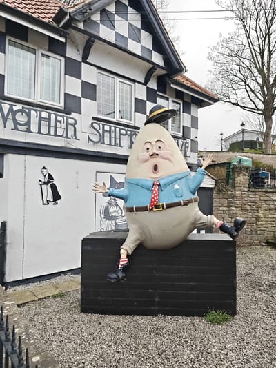 The image depicts a statue of Humpty Dumpty sitting atop a black box-shaped structure in front of the Mother Shipton Inn in Knaresborough, United Kingdom. Humpty Dumpty is wearing a blue shirt, a red polka-dotted tie, beige trousers, and black shoes with striped socks. A hat is precariously balanced on his head. The building behind the statue has a black and white checkered design and lettering which reads "Mother Shipton". To the left of the statue, there is a black and white image of Mother Shipton on the wall. The ground is covered in gravel. A stone wall and trees are visible in the background.