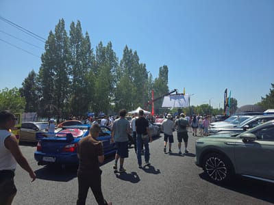 The image captures a sunny outdoor event, likely a car show, in Oskemen, Kazakhstan. A variety of vehicles, including a blue Subaru WRX STI with a prominent spoiler and a green SUV, are parked on an asphalt surface. Several people are walking around, some are looking at the cars, and others are engaged in conversation. In the background, there are trees and banners, suggesting a festive atmosphere. The sky is clear and blue, indicating good weather conditions for the event. The presence of power lines and trees suggests the event is taking place in an open area, possibly a park or a large parking lot within the city.