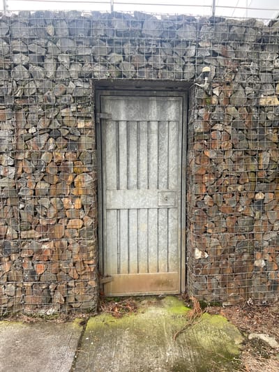 The image depicts a weathered metal door set into a gabion wall. The gabion wall, constructed from wire mesh cages filled with stones, forms the primary structure. The stones are varied in size and color, ranging from dark grey to reddish-brown. The door is grey, with vertical paneling and a horizontal reinforcement bar. A small, circular lock mechanism is visible on the right side of the door. The ground in front of the door is concrete, partially covered with moss and debris such as dry leaves and twigs. The overall scene appears to be outdoors, with a slightly overcast sky visible at the top of the frame. The style of the gabion wall suggests it may be part of a retaining wall or a decorative outdoor feature. No people are present in the image. The context of Balbriggan, Ireland, would imply this is an outdoor structure within a residential, commercial, or public space in that area, possibly a garden boundary, an entrance to a utility area, or part of a landscape design.