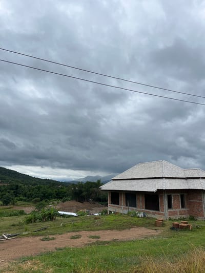 A cloudy, overcast day looms over a rural scene in Ban Mae Na Chalong, Thailand. In the foreground, a dirt path winds through tall, green grass. To the right, several cut tree stumps are scattered on the ground. A partially constructed house, made of red brick with a grey tiled roof, stands prominently in the middle ground. The house has large, dark openings where windows and doors will be installed. Behind the house, a construction site is visible, with piles of dirt, scaffolding, and what appears to be a yellow excavator. The background is dominated by lush green hills and mountains covered in dense trees, disappearing into the hazy sky. Two power lines are stretched across the upper portion of the frame, cutting through the dramatic cloud formations. The overall impression is one of a serene, undeveloped landscape under a moody sky, with ongoing construction indicating future development.