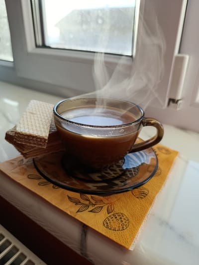 A close-up shot captures a steaming cup of coffee placed on a windowsill. The coffee is in a clear glass mug, revealing its rich brown color. Wisps of steam rise from the surface, indicating it's freshly brewed and hot. Next to the mug, a stack of wafer cookies is neatly arranged. The cup and cookies rest on a patterned orange napkin, which in turn sits on a dark glass saucer. The setting is a windowsill, with a window in the background offering a blurred view of the outdoors. Sunlight streams in, creating reflections on the glass and highlighting the steam. A white radiator is partially visible at the bottom of the frame. The overall impression is one of a cozy, quiet morning, possibly in Pănășești, Moldova, where one might enjoy a warm beverage and a sweet treat. There is no visible text in the image.