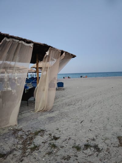 The image depicts a beach scene at Limani Litochorou, Greece, with a focus on an open-air dining or lounge area covered by a thatched roof and draped with sheer curtains. Several tables and chairs are set up beneath umbrellas, with blue lounge chairs visible in the background. The beach itself is sandy and appears relatively clean, with sparse vegetation and debris in the foreground. In the distance, the calm blue sea meets the horizon under a clear sky. A few people are scattered on the beach and near the water, some sitting and others standing, suggesting a leisurely atmosphere. The lighting suggests it is either early morning or late evening, with the sky a pale blue.
