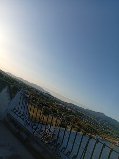 The image depicts a daytime scene in Paiania Municipal Unit, Greece, as viewed from an elevated position, likely a rooftop or balcony. The sky is a gradient of light blue to darker blue. A distant contrail is visible in the upper left quadrant. The foreground features a decorative wrought-iron railing atop a concrete structure. Beyond the railing, a landscape extends, encompassing a mix of vegetation, including trees and scrub, and buildings that are scattered across the terrain. Distant mountain ranges define the horizon. No people are visible in the image.