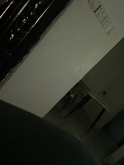 A dimly lit room features a white wall with light switches on the upper left. Next to the wall is a table, also white, with dark-colored legs. A clear bottle is sitting on the table. In the foreground, a large, out-of-focus object obstructs the view. The composition is at an angle, creating a slightly disorienting perspective.
