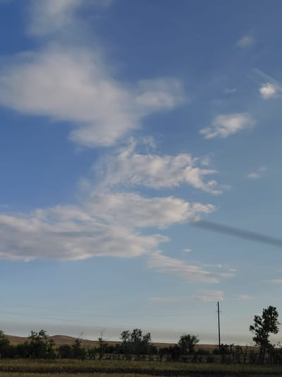 The image captures a wide-angle view of a landscape under a partly cloudy blue sky. In the foreground, there is a field with dry grass and scattered dark soil. A line of trees and shrubs runs across the middle ground, with some appearing bare and others with sparse foliage. A tall, dark utility pole stands among the vegetation on the right side of the frame, with power lines extending horizontally across the scene. In the distance, rolling hills create a soft, undulating horizon. The sky is populated with various white and light gray clouds, some wispy and others more substantial. A faint, dark shadow is cast across the sky from the upper right to the mid-left portion of the image, possibly from an aircraft or a large cloud formation not fully visible. The overall scene suggests an open, rural environment with signs of utility infrastructure. The location is identified as Молодежный, Kazakhstan.