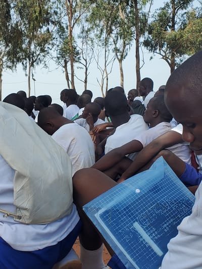 In the city of Barakin Rafin Gora, Nigeria, a group of young people, mostly boys, are gathered outdoors under tall trees with sparse foliage against a clear sky. They are dressed in white tops, with some wearing blue shorts. The individuals are in close proximity, suggesting a communal gathering or event. The foreground is dominated by a close-up of a person in a white t-shirt and blue shorts, holding a blue transparent plastic folder. Another person's head and shoulder, also in a white top, are visible to the left. The folder displays text which includes "CLEAR BAG" and other smaller inscriptions that are partially legible, mentioning "School of Information and Communication" and "Business College," hinting at an educational context. The overall scene appears to be one of a gathering for a school-related activity or event.