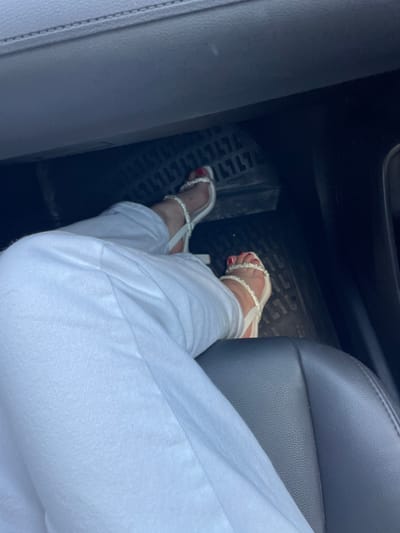 This media file shows a person's legs and feet inside a vehicle. The person is wearing white pants and white jeweled sandals. Their toenails are painted red. The vehicle appears to be a car, with a dark leather seat visible and a black floor mat with a textured pattern. The scene suggests the person is likely a driver or passenger. The lighting indicates it is daytime. The presence of a car interior with pedals and seating suggests an indoor, stationary setting, possibly a parking area or street. There is no visible text in the image.