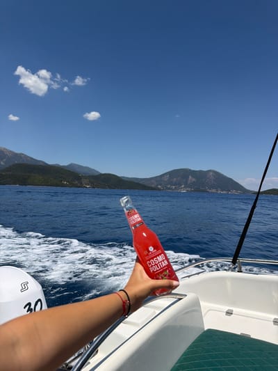 The image shows a person holding a bottle of Cosmoopolitan cocktail on a boat sailing in the sea. The boat is white with a black fishing rod attached to it. In the background, there are green mountains and blue sky with clouds. The water is blue and choppy, with white foam created by the boat's movement. The person is wearing a red string bracelet on their left wrist. The location is the sea near Spartochori, Greece. The scene depicts a leisure activity on a sunny day.