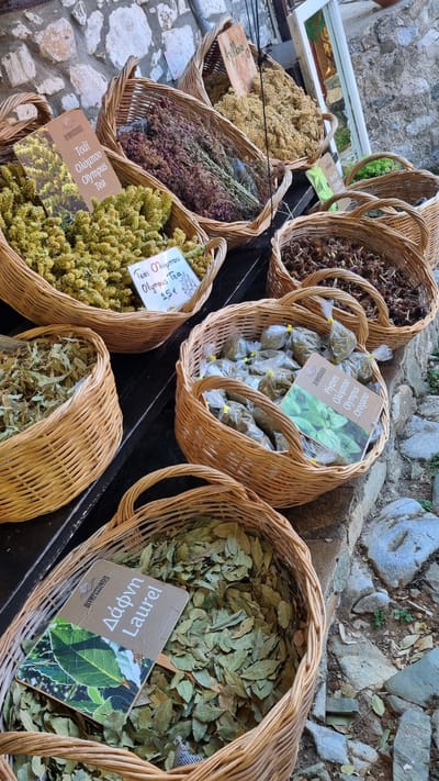 This is a close-up, top-down shot of several woven baskets filled with dried herbs and spices, likely at an outdoor market stall. The baskets are arranged in a tiered fashion, with some spilling over the edges. The herbs vary in color and texture, ranging from fine, light yellow clusters to darker, more stringy purple and brown varieties, as well as a basket full of green leaves.
One basket prominently displays small, individually packaged bags of dried herbs, with a label indicating "Olympus Oregano". Another basket contains a large quantity of dried green leaves, identified by a label as "Laurel". In the foreground, a basket is filled with small, light green, flower-like dried herbs, with a label reading "Olympus Tea" and a price of "1.5€". The ground beneath the baskets is paved with irregular stones.
The scene suggests a rustic, traditional setting, consistent with a village market in Greece, as indicated by the Greek text on the labels. The lighting suggests it is daytime, and the overall impression is one of abundance and natural products. The text visible on the labels includes product names like "Oregano", "Laurel", and "Olympus Tea", along with brand information such as "Olympussenses".