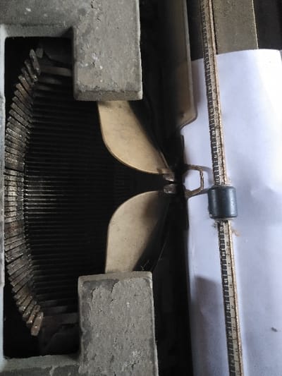 A close-up, vertical view depicts the internal components of an aged mechanical typewriter. On the left, a curved assembly of numerous metal type bars is visible, largely in shadow. Centrally, a blank sheet of white paper is positioned against two curved, pale-colored paper guides. To the right of the paper, a vertical metal ruler with metric markings, numbered from 0 to 55 in increments of 5, is visible, accompanied by a dark, cylindrical paper-holding component. Two prominent, grey, rectangular structural elements of the typewriter frame appear in the foreground, exhibiting chipped surfaces, accumulated dust, and grime, with faint cobwebs present in crevices. The scene is located in the city of Жібек Жолы, Kazakhstan.