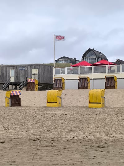 The image depicts a cloudy day at a beach, likely in or near Bergen, Netherlands. In the foreground, several yellow beach chairs, a common sight in Dutch coastal towns, are arranged on the sand. Behind them, a low-lying wooden building with stairs and a sign reading "VEEHUIZ" is visible. Further back, a row of beach chairs with striped awnings are positioned in front of a building with a distinctive arched roof. Red umbrellas with a Coca-Cola logo are also present in this area. A white flag with the red "TEXELS" logo flies from a flagpole against the overcast sky. The overall scene conveys a quiet, possibly off-season, beach atmosphere.