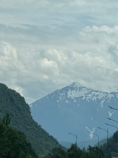 The image shows a panoramic view of a mountainous landscape with a partially snow-capped peak in the distance. The foreground features a steep, tree-covered mountainside on the left, with dark green foliage. In the middle ground, the imposing mountain range dominates the scene, its slopes streaked with snow. The sky above is filled with a mix of white and grey clouds, suggesting overcast conditions. Several streetlights with curved poles are visible in the lower right portion of the image, indicating the presence of a road or urban infrastructure near the natural landscape. The overall impression is one of natural beauty and a blend of rugged wilderness with hints of human development. The location is identified as Kepsha, Russia, a village in the Lazarevsky District of Sochi, known for its mountainous terrain and proximity to the Black Sea.