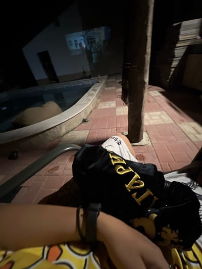 The image captures an outdoor night scene in Debelt, Bulgaria, presenting a low-angle perspective from a reclining individual. The foreground features a gray armrest, likely from a lounge chair, and the person's lap, covered by a black garment with large gold lettering ("KAPE" or "LAPE") over a yellow and white patterned fabric. The individual wears white shorts with dark text visible on the left thigh, and a black wristband or smartwatch is on their left wrist.

The immediate environment consists of a patio paved with reddish-brown and lighter rectangular tiles, showing some wet spots. To the left, a swimming pool with a light-colored coping contains dark water, with a tan, rounded object partially submerged. In the background, an image, appearing to be an interior room with windows, is projected onto the white exterior wall of a building, which also has a dark doorway. A thick, dark pillar or post with visible metal attachments stands vertically in the midground, towards the right. Further right, stacked flat objects are faintly discernible in the shadows. The overall lighting is dim, primarily from the projection and ambient sources.