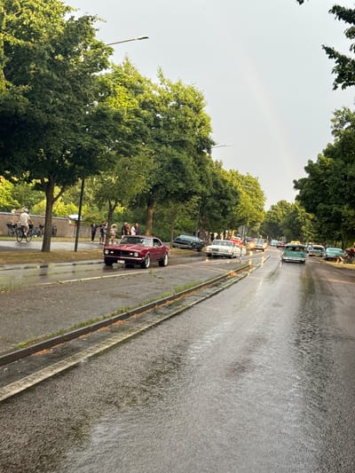 The image captures a street scene in Falkenberg, Sweden, under overcast skies with a faint rainbow visible. The road is wet, indicating recent or ongoing rain, with visible puddles and reflections. A procession of vintage cars is driving down the street. In the foreground, a dark red classic car, possibly a Chevrolet Camaro, is the most prominent vehicle. Further down the road, other classic cars, including a white one and a teal-colored one, are visible. Several people are gathered on the sidewalks and near the cars, some watching the vehicles, while others appear to be enjoying the event. Trees line the street, their foliage lush and green. A street light is also visible. On the left side of the road, a person on a bicycle is seen near parked bicycles. The overall atmosphere suggests a car show or a classic car rally taking place in a residential or tree-lined urban area.