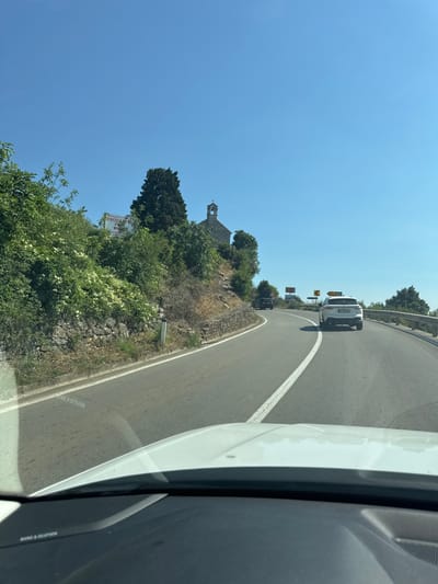 The image depicts a view from the windshield of a white car driving on a winding road in Blizikuće, Montenegro. The road curves to the right, with a white line marking the lane. On the left side of the road, a steep, rocky embankment is covered with dense green vegetation, including flowering bushes. A small stone building with a bell tower, possibly a church, is visible atop the embankment, nestled among trees. A white sign with red lettering is also present on the embankment. Further down the road, a white SUV is in front of the viewer's car, and another dark SUV is ahead of it. The sky is clear blue. The foreground shows the dashboard and part of the hood of the car from which the image is taken.