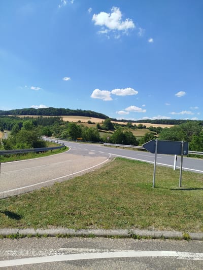 The image shows a rural intersection in the city of Bad Mergentheim, Germany, on a clear day with a blue sky and scattered white clouds. The intersection features two asphalt roads meeting at an angle, with white lane markings. A metal guardrail runs along the side of the road. In the foreground, there is a patch of green grass with a concrete curb. A grey directional sign with an arrow pointing left is mounted on two poles to the right of the road. The background consists of rolling hills covered in trees and fields of golden crops, suggesting agricultural land. There are no people or vehicles visible in the image.