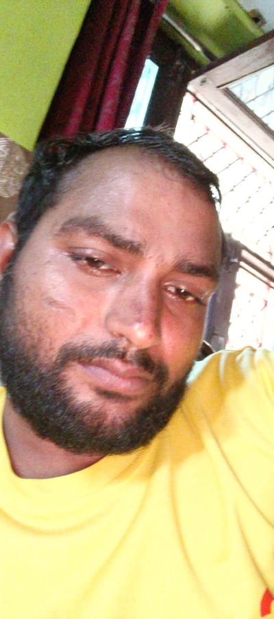 The image shows a close-up of a man's face and upper torso. The man has short black hair, a dark beard and mustache, and is wearing a yellow t-shirt. He is looking directly at the camera with a neutral expression. The man is likely sitting in a chair or on a couch, with his body turned slightly to the right. The image is taken from a low angle. The location based on the coordinates is in the city of Noida, India.