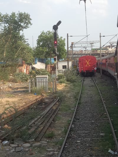 The image shows a railway scene in Bhopal, India. A red train car with a large yellow "X" is coupled to another car on a track that curves away from the viewer. Overhead electrical lines and signal equipment are visible. A signal light is showing red. Adjacent to the tracks are ballast, vegetation, and discarded materials. A small white building and partial views of other structures are visible behind the tracks. Discarded rails are stacked in the foreground.
