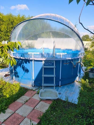 This is a medium shot, exterior, eye-level, daylight photograph of a blue, round, above-ground swimming pool with a clear, domed canopy. The pool is surrounded by green grass, weeds, and some flowering plants. A decorative pathway of square, red and white tiles leads up to the pool. The pool has a silver ladder leading into it, and a filter pump and hose are visible on the right side of the image. The sky is blue with a few scattered white clouds. The scene is set in a backyard garden in the village of Vyazki, Russia. There are no people or animals present.