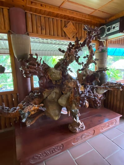 The image depicts an intricate wooden sculpture of a dragon, a mythical creature, displayed on a dark wooden table within a room featuring a rustic, wooden interior. The sculpture is highly detailed, showcasing the dragon's head with an open mouth, scales, and claws, intertwined with branches and other natural elements. The room has large windows with a view of lush greenery, suggesting an outdoor setting visible through the glass. The presence of a speaker system mounted on the wall and decorative baskets hints at a domestic or recreational space. The lighting appears natural, filtering in through the windows. The specific location in Thôn 2, Vietnam, indicates a rural or semi-rural environment.