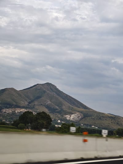 The image captures a scenic view of a mountain range under a cloudy sky. In the foreground, a concrete barrier, likely part of a highway, is visible with a blurred effect, indicating movement. Several rectangular signs with white and black markings are mounted on poles along the barrier. To the left, there are three round hay bales in a field, bordered by green grass and trees. In the middle ground, a cluster of mature trees stands tall, separating the field from a distant mountainside dotted with scattered houses and buildings. The mountain itself is covered in vegetation and features exposed rock formations, particularly on its slopes. The sky is filled with a mixture of gray and white clouds, suggesting an overcast day. The location is identified as Bellona, Italy.