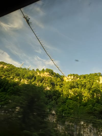 The image, taken with a slightly rotated perspective, showcases a dramatic landscape, likely in the Galitsyno area of Russia, under a clear, bright sky dotted with wispy white clouds. A long, slender suspension bridge-like structure or aerial pathway spans horizontally across the frame, stretching over what appears to be a valley or gorge.

On the right side of the image, a large, steep mountain or hill dominates, covered in lush, vibrant green foliage. Sections of the mountain are bathed in a warm, golden light, suggesting it is daytime, possibly morning or late afternoon. Along the aerial structure, a distinct, cylindrical cabin or gondola can be seen, indicating its function as an aerial tramway or a similar transport system for passengers or goods.

In the distance, atop the mountain, several tall, white, lattice-framework towers, likely electricity pylons, stand against the sky. Thin, dark power lines run vertically along the far right edge of the frame, parallel to the mountainous terrain. The bottom-left corner of the image shows a dark, blurry element, consistent with a reflection or interior part of a vehicle, suggesting the photograph was captured from within a moving transport. No visible text is present in the image.