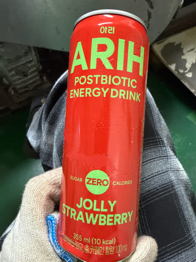 A red beverage can, branded "ARIH POSTBIOTIC ENERGY DRINK" in green lettering, is held by a person wearing a beige glove. The can is slightly damp with condensation. Below the brand name, "SUGAR ZERO CALORIES" is visible, also in green. Further down, "JOLLY STRAWBERRY" is written, followed by "355 ml (10 kcal)". The background is out of focus, showing a dark, textured surface and a green floor. This scene is located in Hwaseong-si, South Korea.